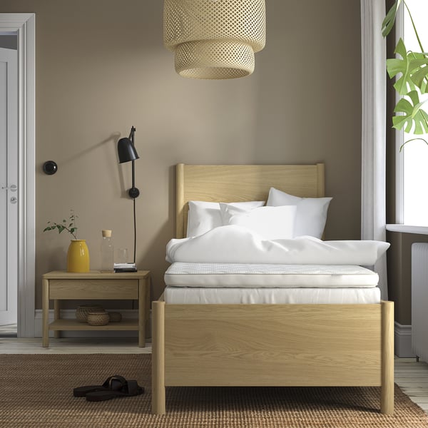 Minimalist bed with wooden headboard, white linens, hanging light, side table with lamp, and yellow vase. Bed is larger than side table.
