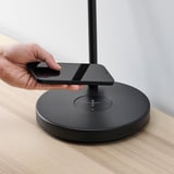 Hand placing smartphone on wireless charging pad atop circular base, beside modern lamp on table.