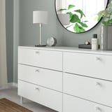 White dresser with six drawers, each having round knobs for easy gripping. Topped with a round mirror, lamp, and decorative items.