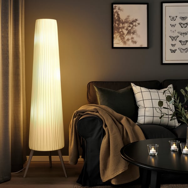 ÖKENSAND floor lamp beside couch, emitting soft light, nearly reaching ceiling.