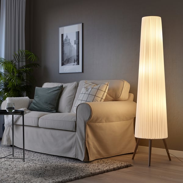 Living room with beige floor lamp, illuminating a cozy space. Gray sofa with green pillows, side table, plant, and curtains complete the modern decor.