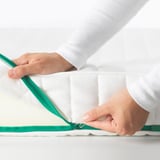 In hands are zipping a green zipper on a white mattress, demonstrating its easy-to-clean, machine-washable cover.