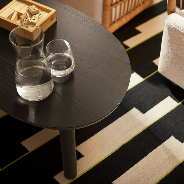 An öträsk round coffee table in black with a glass jug and cups. The table is placed on a black and beige striped rug, slightly smaller than the rug.