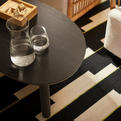 An öträsk round coffee table in black with a glass jug and cups. The table is placed on a black and beige striped rug, slightly smaller than the rug.