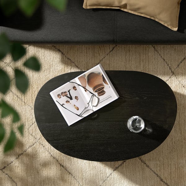 Dark brown ÖRTRÄSK coffee table with items on it, near black couch and green plant.