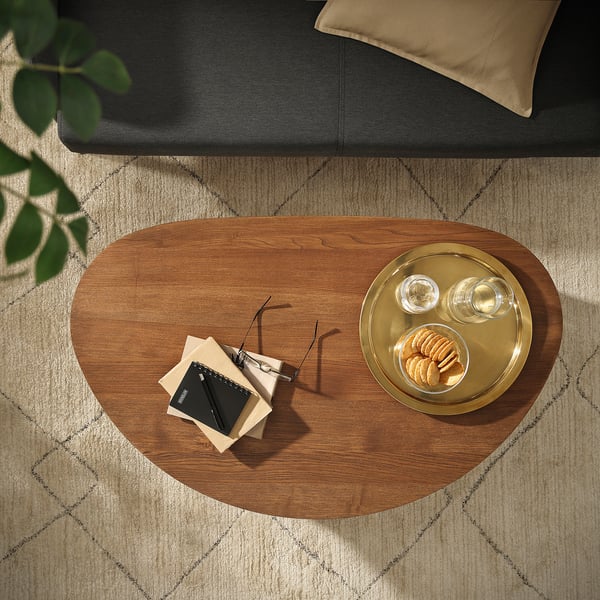 ÖRTRÄSK coffee table with natural wood, gold platters, remote, papers. Black sofa with beige cushion contrast.