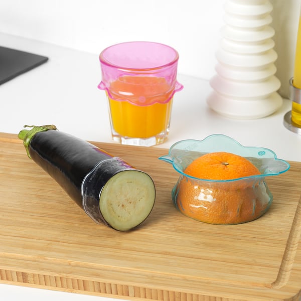 Wooden cutting board with half-cut eggplant and an orange in a silicone cover next to a glass of juice and stackable vases.