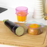 Wooden cutting board with half-cut eggplant and an orange in a silicone cover next to a glass of juice and stackable vases.