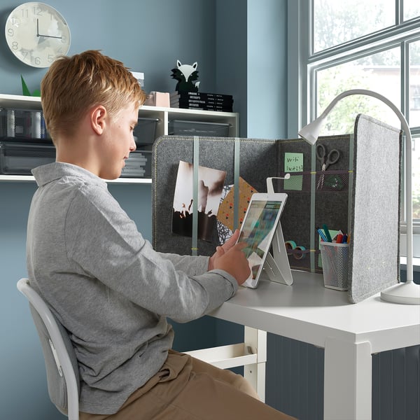Boy sitting at white desk using tablet. Desk has gray divider for privacy and sound dampening; holds notes and supplies.