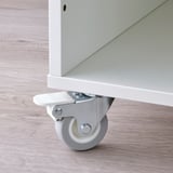 Close-up of a white caster wheel attached to a white wooden furniture leg on a light wooden floor.