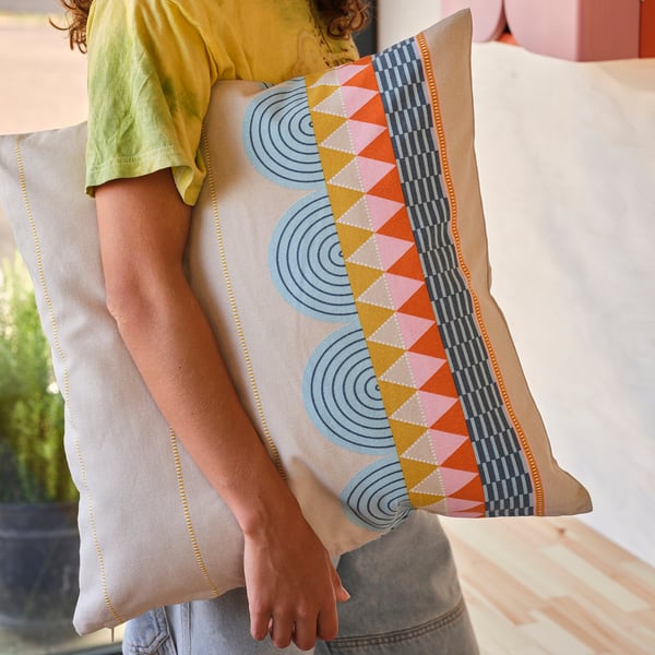 Person holding OMMJÄNGE cushion with geometric patterns on bed.