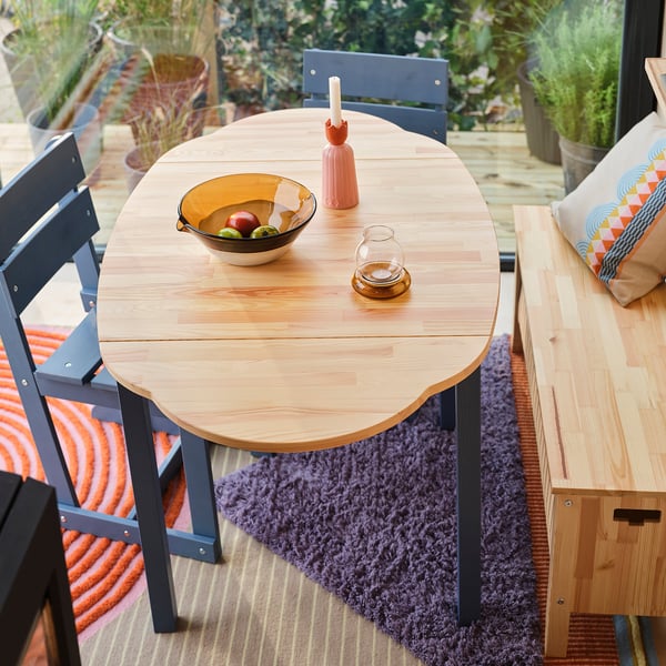 Blue-legged pine table with foldable leaves on purple rug. Holds fruits, vase, glass, with nearby bench.