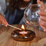 Person lights a tealight candle using a match inside a glass holder on a wooden table.