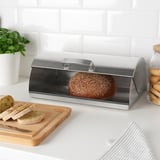 Stainless steel bread box with loaf, sliced bread, cookies, and plant on white countertop.