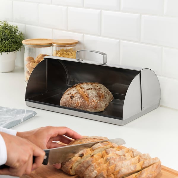 Person slicing bread with a knife. A metallic bread box with a loaf inside sits on a white countertop.