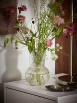 A clear glass PÅDRAG vase with a round, ribbed design, holding a bouquet of pink and white flowers with green stems and leaves.