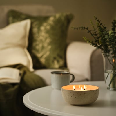 A lit PÄRLHÖNA scented candle in a speckled stoneware bowl sits on a coffee table, smaller than an enamel mug and a vase with greenery.