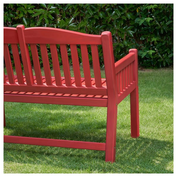 A red PÄRONHOLMEN bench made of acacia wood. The bench has a curved back and straight slats, designed for outdoor use, placed on a grassy lawn.