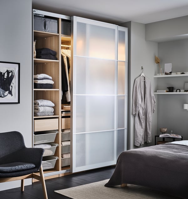 A modern bedroom with a PAX wardrobe in white, featuring glass doors, organized shelves, baskets, and a grey bed.