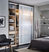 A modern bedroom with a PAX wardrobe in white, featuring glass doors, organized shelves, baskets, and a grey bed.