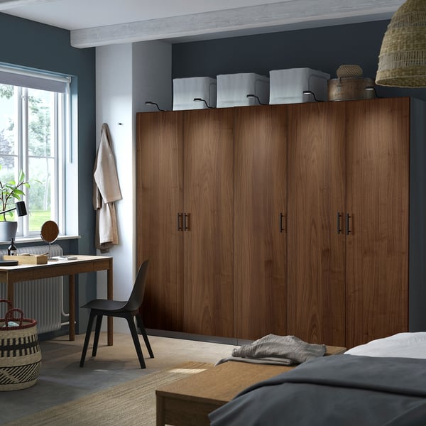 Large wooden PAX wardrobe with multiple doors stands beside window and desk. Its size dominates the room, storing clothes and items efficiently.
