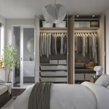 Modern bedroom with open wardrobe showing clothes, accessories, and lit shelves. Soft-close hinges visible, integrated lighting on shelves.