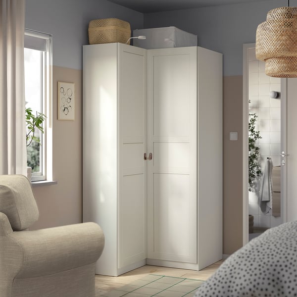 A white corner PAX wardrobe with double doors and shelves. It fits perfectly in a living room with beige furniture and a window.