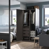Modern bedroom with black PAX wardrobe, gray armchair, and white bed. Clothes hang inside open wardrobe, gray wall, large window.