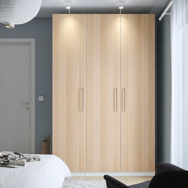 Minimalist bedroom with a tall light wood PAX wardrobe, white bed, and black chair against a dark wall.