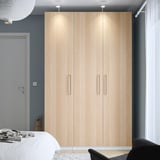 Minimalist bedroom with a tall light wood PAX wardrobe, white bed, and black chair against a dark wall.
