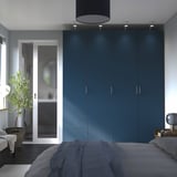 Modern blue bedroom with large PAX wardrobes. Dark blue bed linens. Tall, sleek cabinets against wall. Glass door to bathroom. Plant on left.