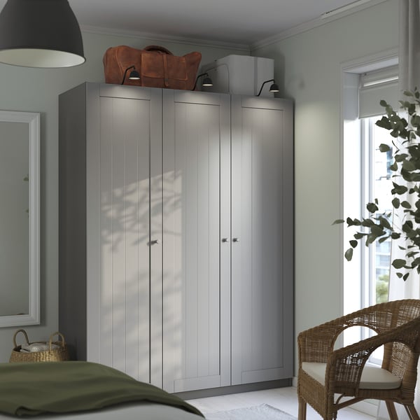 Gray ikea PAX wardrobe with two doors, between a window and wall. Topped with a bag and box, beside a wicker chair with a green blanket.