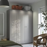 Gray ikea PAX wardrobe with two doors, between a window and wall. Topped with a bag and box, beside a wicker chair with a green blanket.