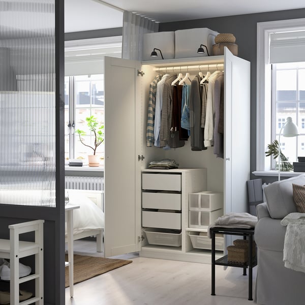 A well-lit wardrobe displaying clothes and drawers, part of an organized bedroom with a view into a cozy living area.
