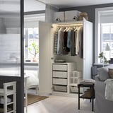 A well-lit wardrobe displaying clothes and drawers, part of an organized bedroom with a view into a cozy living area.