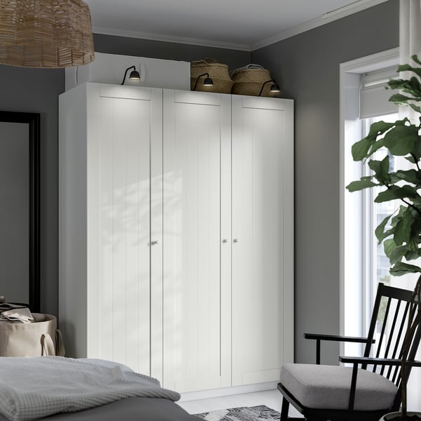 Large white PAX wardrobe stands tall, spanning the room height, flanked by wicker baskets and subtle lighting, near a window with plants.