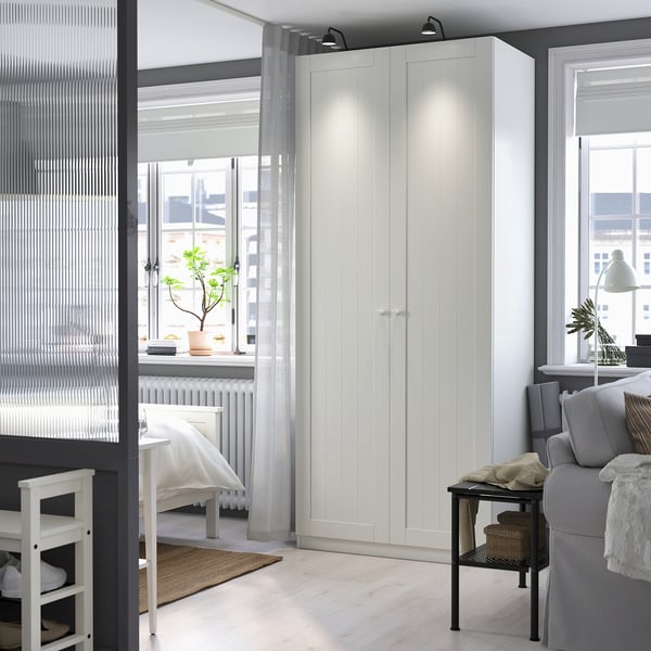 A white PAX wardrobe between window & wall, beside grey sofa, clean & organized.