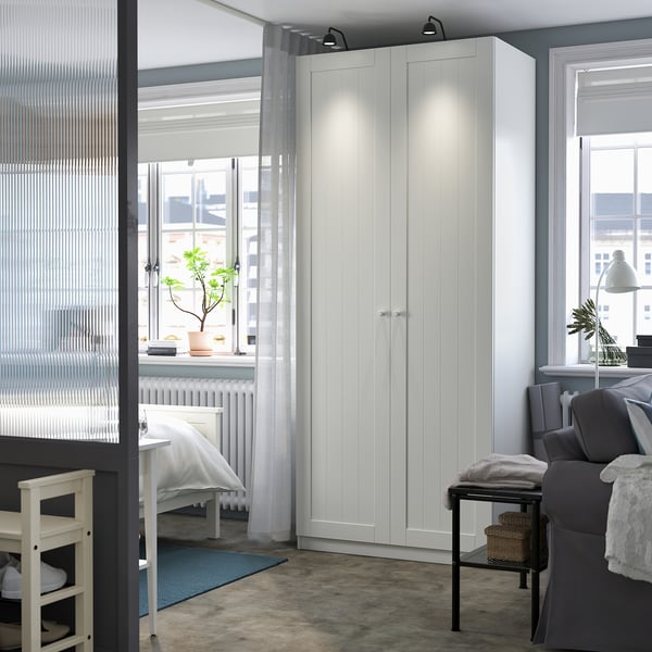 Modern white PAX wardrobe with storage, sleek panels, lighting, surrounded by windows, sofa, rug.