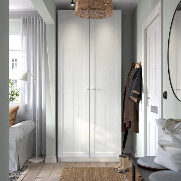 Modern entrance with tall white PAX wardrobe, jacket, boots, mirror, grey sofa, lamp, and window.