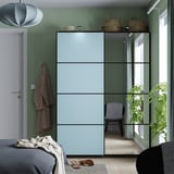 Modern green bedroom with PAX wardrobe, mirror, plants, and hanging coat. Blue bed with grey blanket.