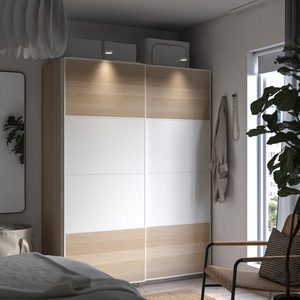 Modern bedroom with PAX wardrobe, light wood and white finish, sliding doors, soft lighting, wicker chair, and large window.