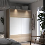 Modern bedroom with PAX wardrobe, light wood and white finish, sliding doors, soft lighting, wicker chair, and large window.