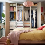 Bedroom with open wardrobe showing clothes in hangers, shelves with boxes, and a bed with yellow bedding.