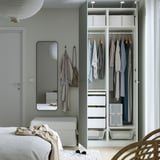 Modern bedroom with open wardrobe showing clothes, drawers, and hanging jackets. Light grey walls, mirror, and white furniture.