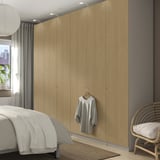 A modern bedroom with a light-colored wooden PAX wardrobe, larger than the nearby white bed and beige chair. It sleek doors and integrated lighting.