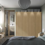 Modern wooden PAX wardrobe with soft-close doors and lighting. Two boxes on top, beside a door, next to a bed and vase.