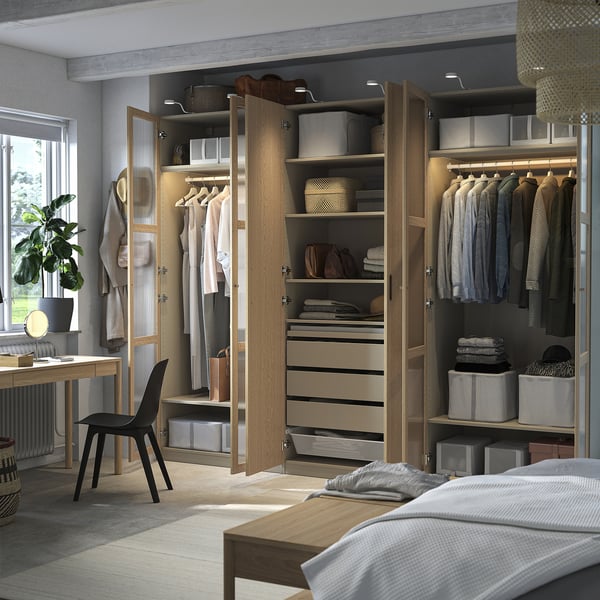 Open wooden wardrobe displaying organized clothes, accessories, and shelves in a bedroom with a desk and chair.