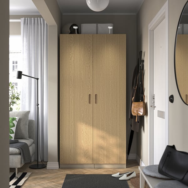 Modern bedroom with tall wooden wardrobe, gray chair, black lamp, and mirror by white door.