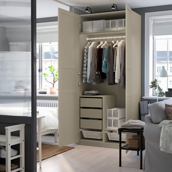 Beige PAX wardrobe with shelves, drawers, and hanging rod. Lit inside, stands in well-lit room with large window and grey chair.