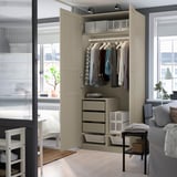 Beige PAX wardrobe with shelves, drawers, and hanging rod. Lit inside, stands in well-lit room with large window and grey chair.
